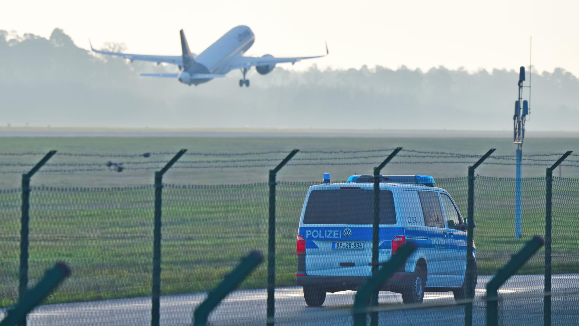 Ein Polizeiauto steht hinter einem Sicherheitszaun auf einem deutschen Flughafen. Im Hintergrund startet ein Flugzeug.