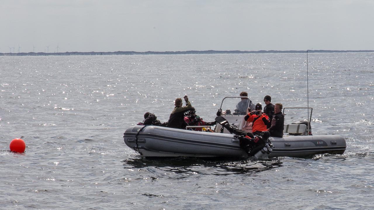 Ein graues Schlauchboot auf dem Meer, an Bord sind rund ein halbes Dutzend Personen. Eine Person trägt eine rote Jacke. Ein Taucher in schwarzem Anzug und großen Schwimmflossen an den Füßen wirft sich rücklings ins Meer. Ein graues Schlauchboot auf dem Meer, an Bord sind rund ein halbes Dutzend Personen. Eine Person trägt eine rote Jacke. Ein Taucher in schwarzem Anzug und großen Schwimmflossen an den Füßen wirft sich rücklings ins Meer.