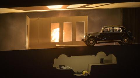 Auf einer Bühne steht ein Auto vor großen fensterartigen Bauten, aus denen viel Licht scheint. Darunter erscheint eine Art Keller, der die gespiegelte Form des Autos auf der Bühne hat.
