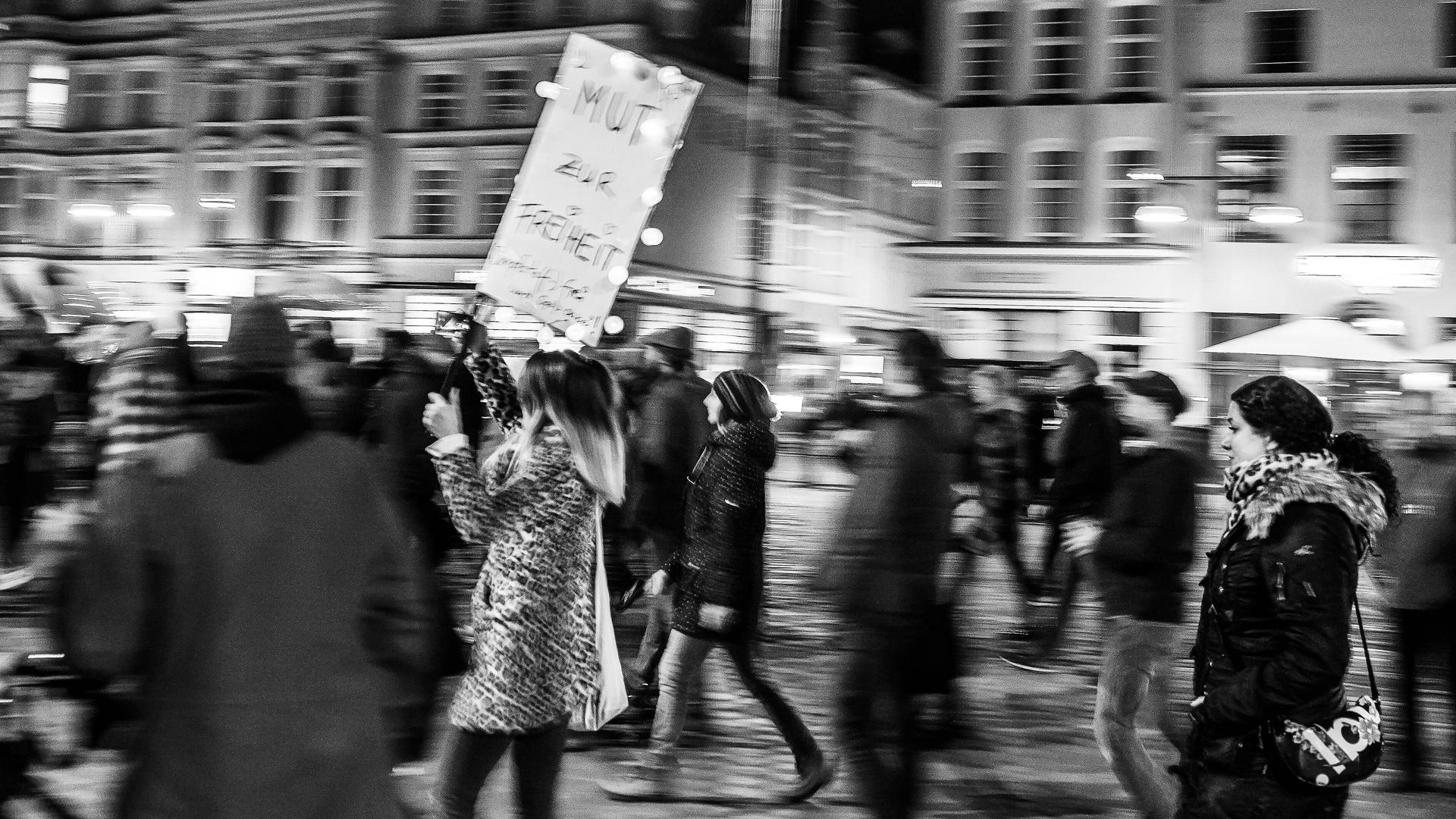 Bewegungsunscharfes Schwarz-Weiß-Foto einer Frau, die auf einer Querdenkerdemo ein Schild mit der Aufschrift "Mut zur Freiheit" trägt.