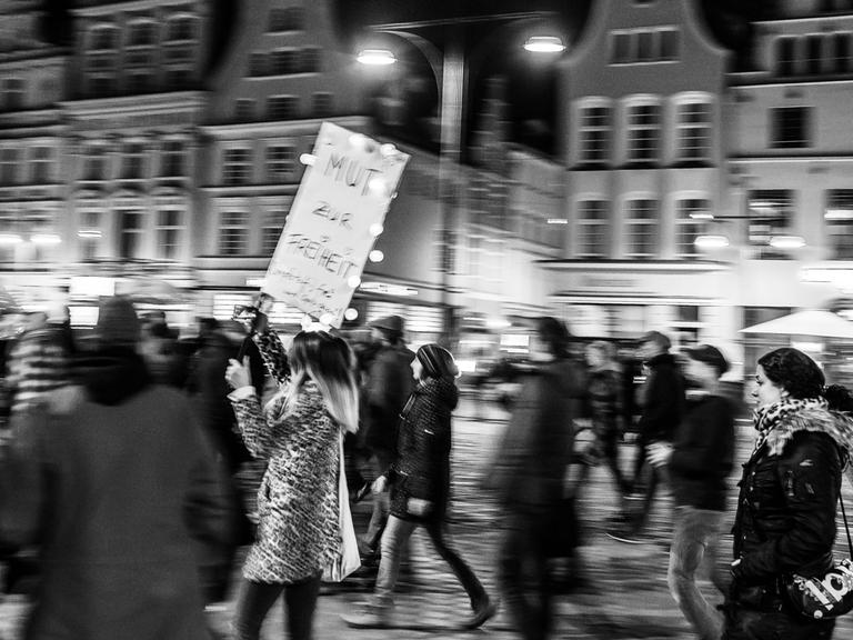 Bewegungsunscharfes Schwarz-Weiß-Foto einer Frau, die auf einer Querdenkerdemo ein Schild mit der Aufschrift "Mut zur Freiheit" trägt.