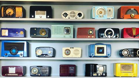 In einem schrankhohen Regal sind ganz unterschiedliche, bunte Radiogeräten einsortiert.