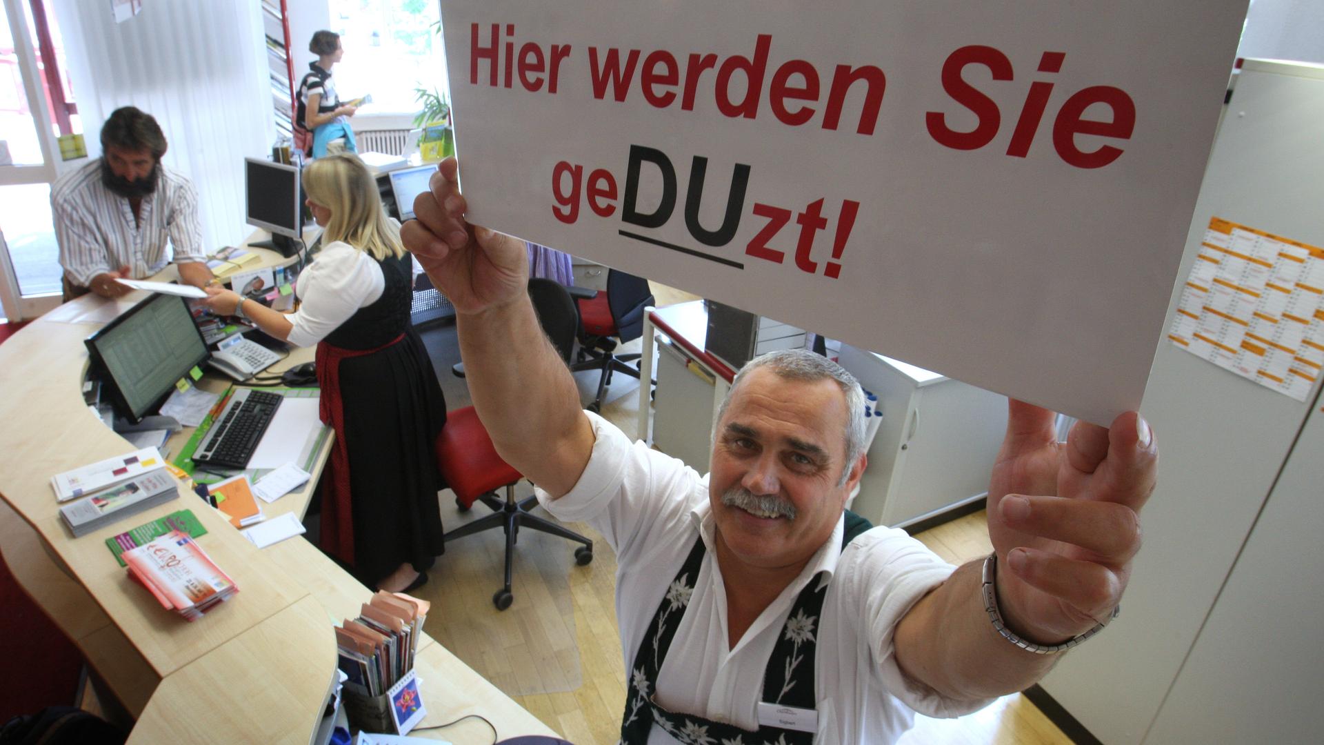 Ein Mitarbeiter der Oberstaufen Tourismus Marketing GmbH hält am "Du"-Schalter im Haus des Gastes in Oberstaufen (Schwaben) ein Schild mit der Aufschrift "Hier werden Sie geDUzt" hoch.