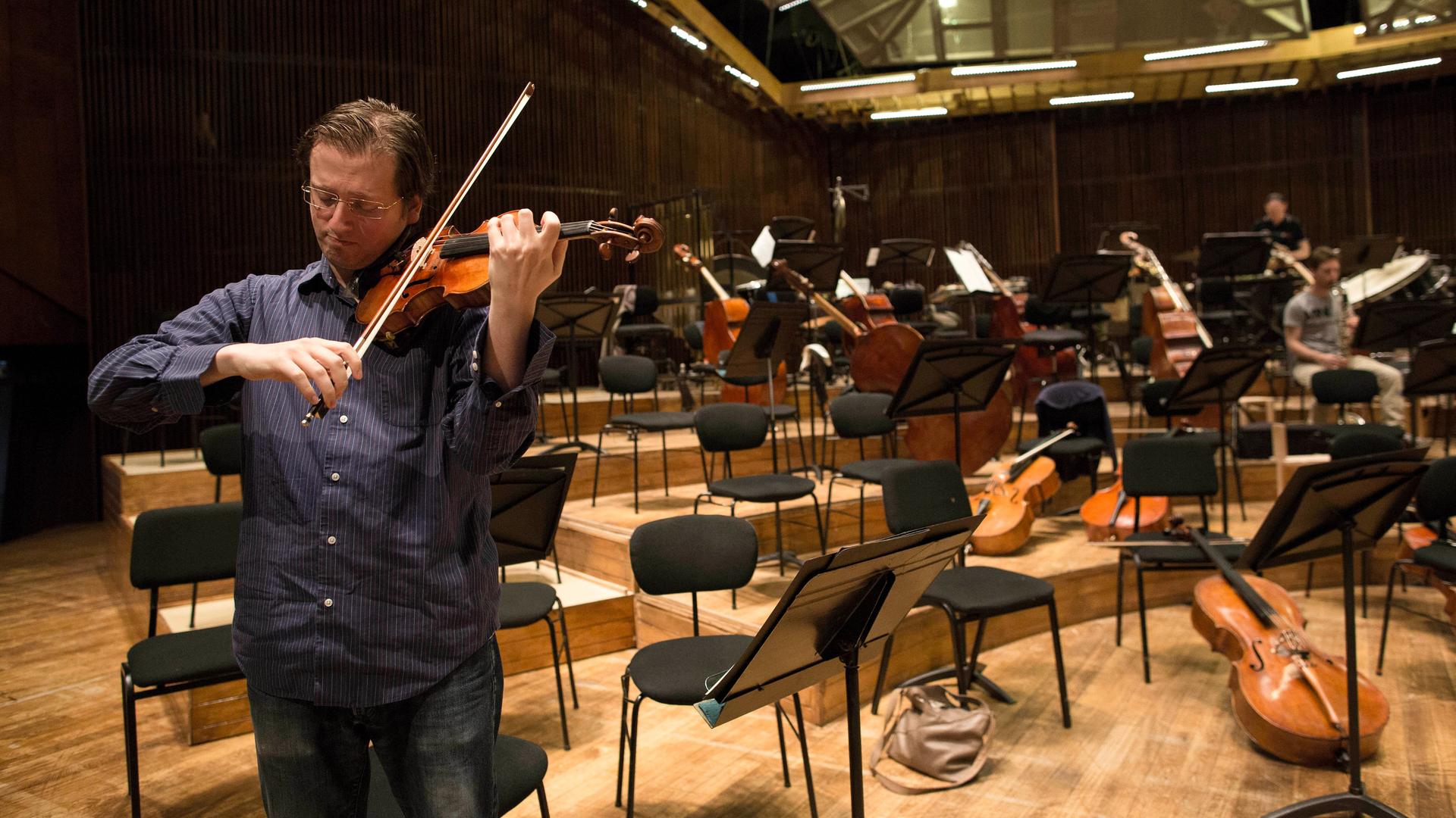 Ein Geiger steht am Rand einer Bühne. Die anderen Plätze des Orchesters sind leer
