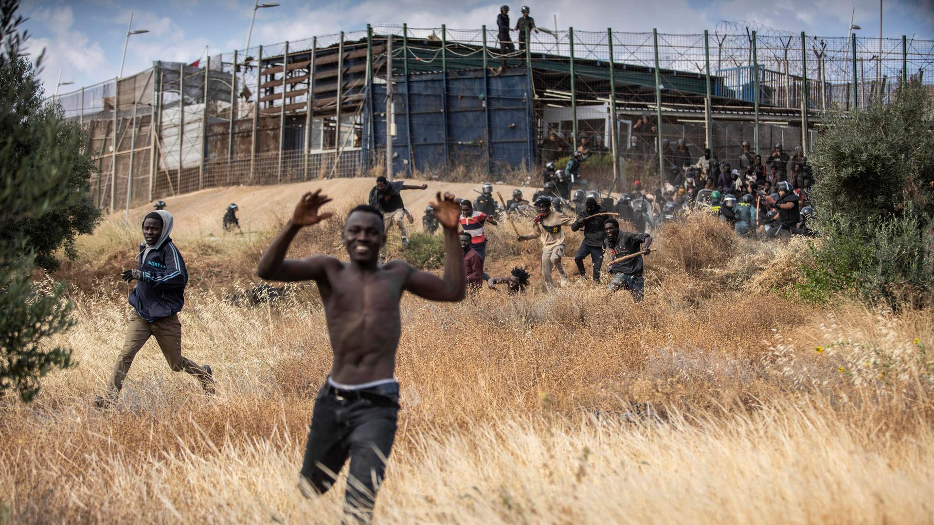 Migranten rennen auf spanischem Boden, nachdem sie den Grenzzaun zwischen Melilla und Marokko überwunden haben. Im Hintergrund ist der Zaun zu sehen, an dem immer noch Migranten versuchen, an den Sicherheitskräften vorbeizukommen.