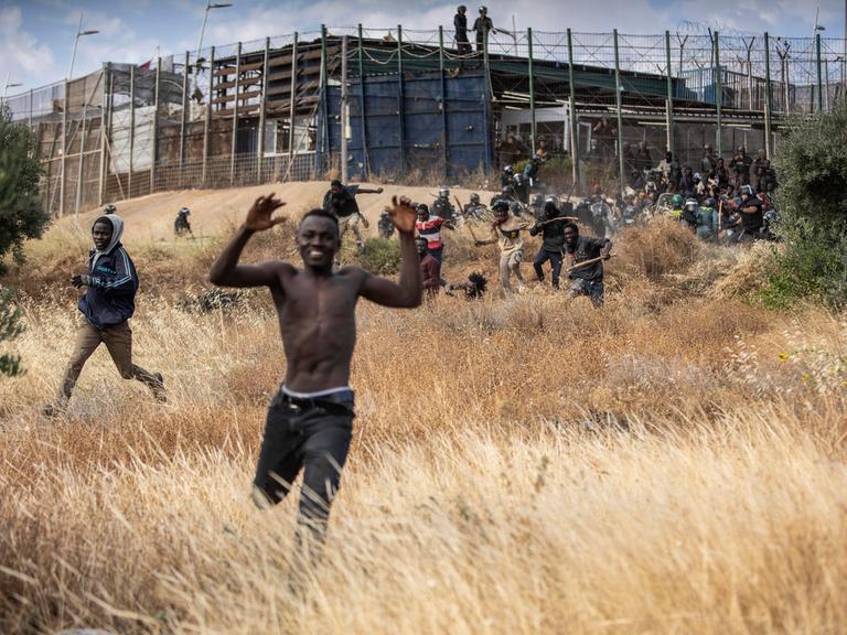 Migranten rennen auf spanischem Boden, nachdem sie den Grenzzaun zwischen Melilla und Marokko überwunden haben. Im Hintergrund ist der Zaun zu sehen, an dem immer noch Migranten versuchen, an den Sicherheitskräften vorbeizukommen.