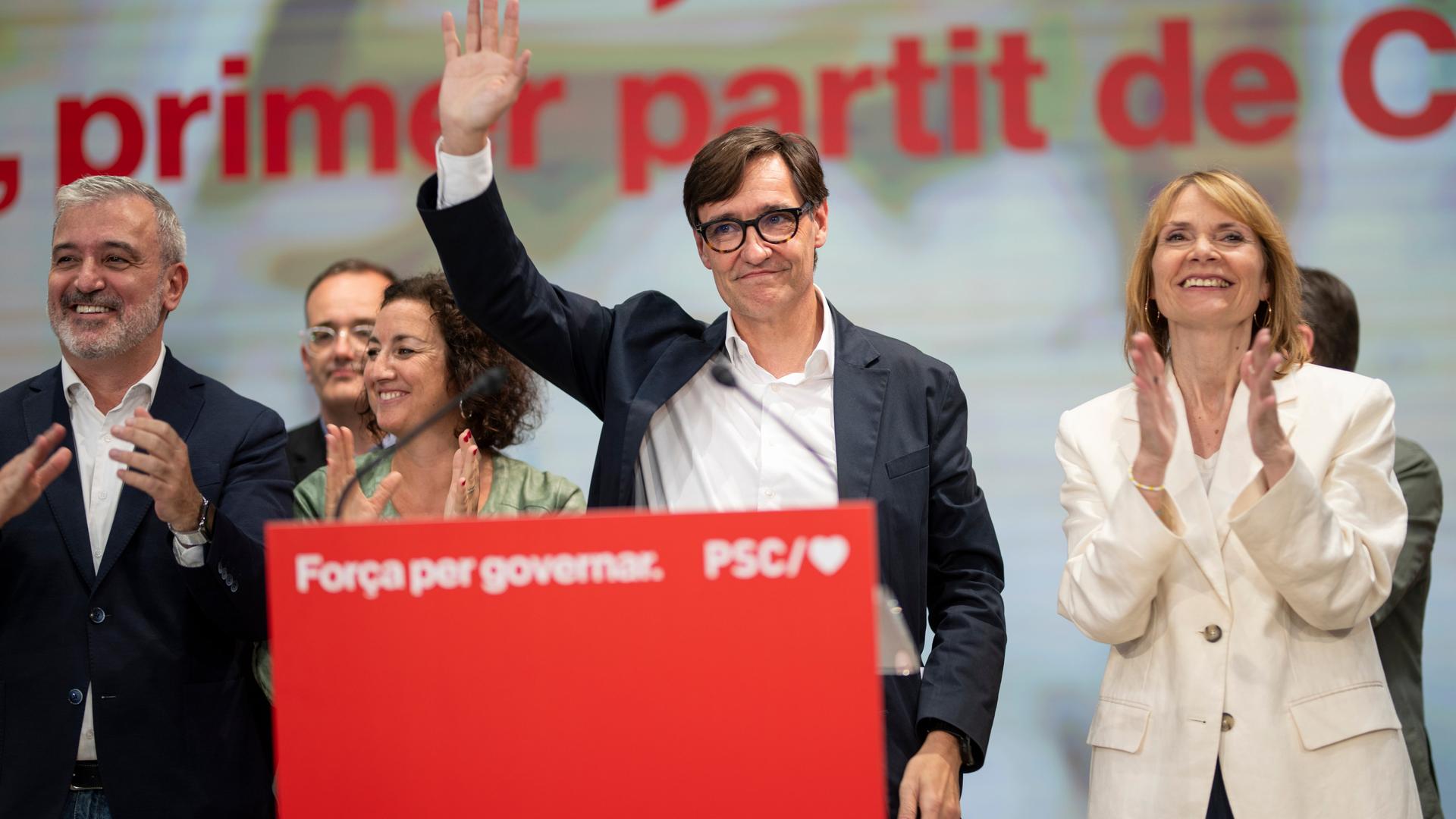 Illa mit Brille und schwarzem Jackett steht auf einer Bühne an einem roten Rednerpult und winlt lächelnd. Daneben klatschen zwei Frauen und drei Männer. Illa mit Brille und schwarzem Jackett steht auf einer Bühne an einem roten Rednerpult und winlt lächelnd. Daneben klatschen zwei Frauen und drei Männer.