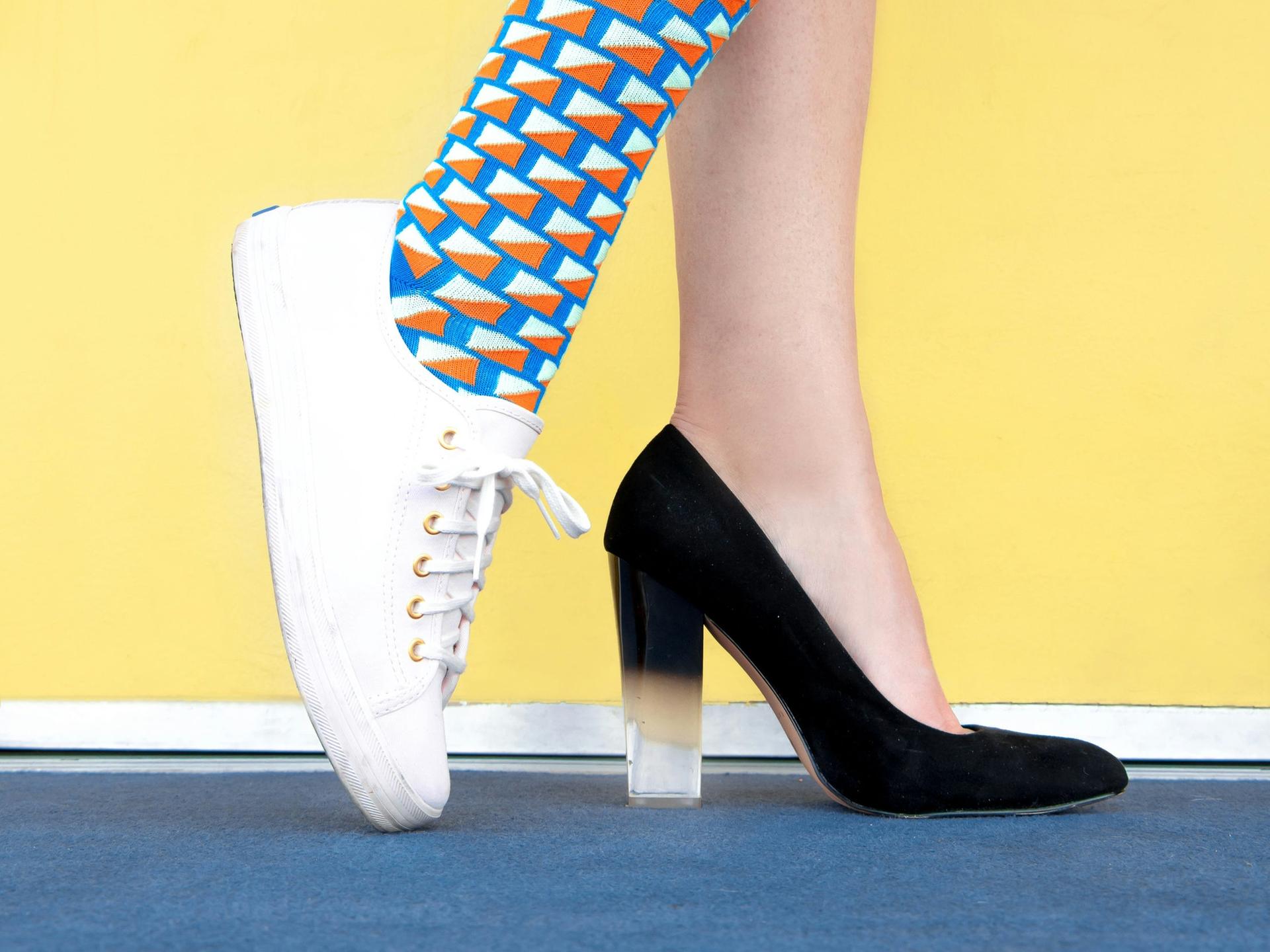 Blick auf die Füße einer Frau, die mit dem einen Fuß in einem schwarzen Pumps steckt und mit dem anderen in bunter Socke in einem weißem Turnschuh.