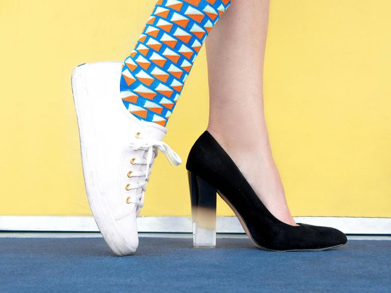 Blick auf die Füße einer Frau, die mit dem einen Fuß in einem schwarzen Pumps steckt und mit dem anderen in bunter Socke in einem weißem Turnschuh.