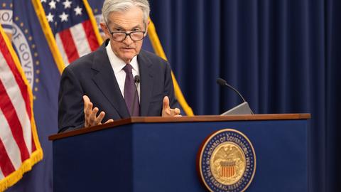 Powell im dunklen Jackett steht an einem Rednerpult mit dem Wappen der Fed. Er spricht und gestikuliert mit der rechten Hand. Dahinter zwei US-Flaggen.