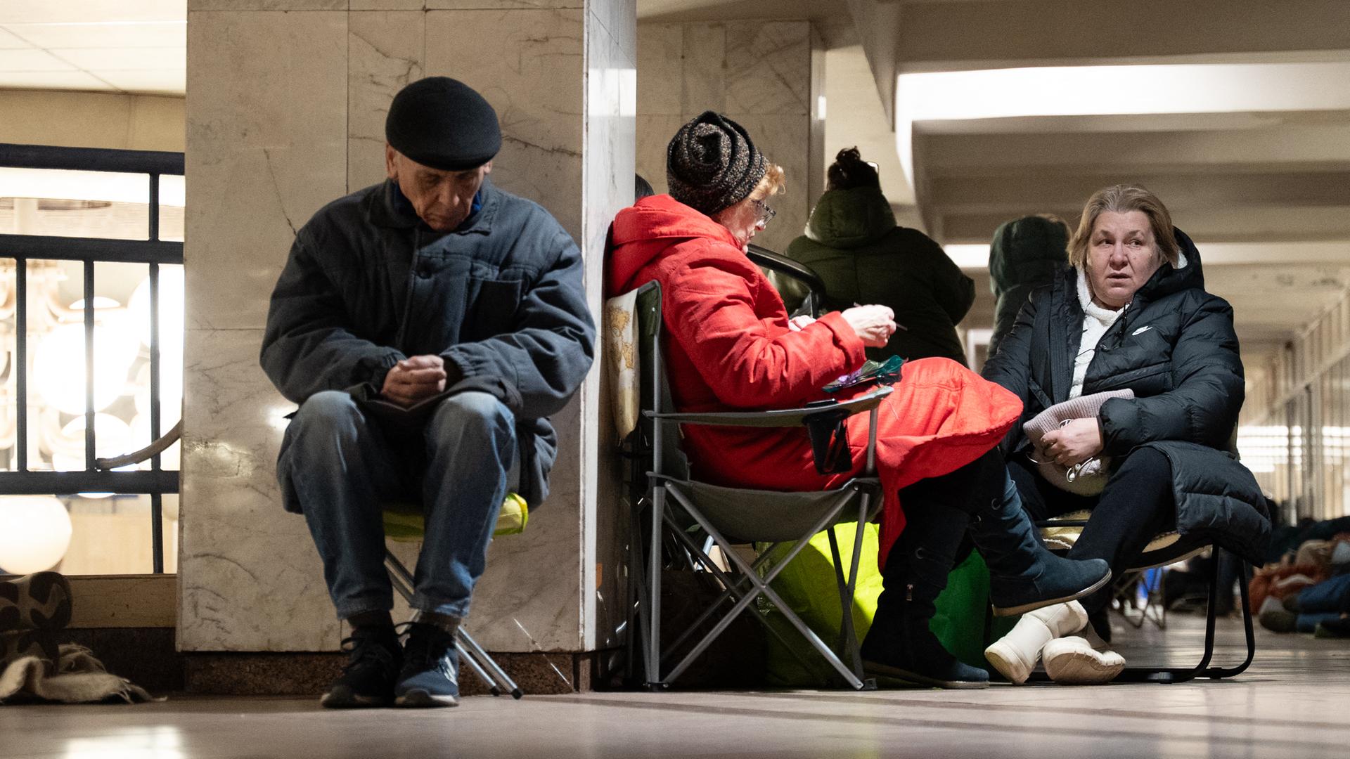Ein älterer Mann sitzt in Winterkleidung auf einem Klappstuhl an eine Wand gelehnt und scheint zu schlafen. Neben ihm zwei Frauen auf Klappstühlen.