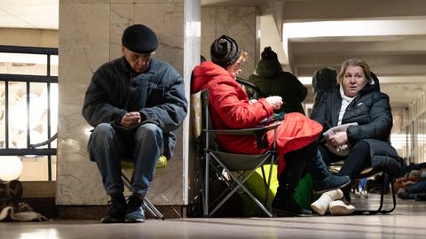 Ein älterer Mann sitzt in Winterkleidung auf einem Klappstuhl an eine Wand gelehnt und scheint zu schlafen. Neben ihm zwei Frauen auf Klappstühlen.
