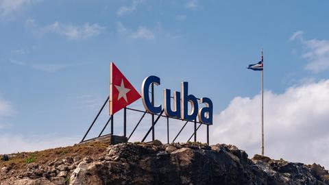 Eine kubanische Fahne an einem Fahnenmast hinter einem großen Schriftzug "Cuba" auf einem Felsen