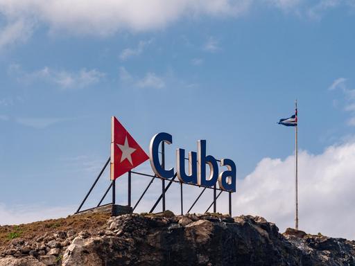 Eine kubanische Fahne an einem Fahnenmast hinter einem großen Schriftzug "Cuba" auf einem Felsen
