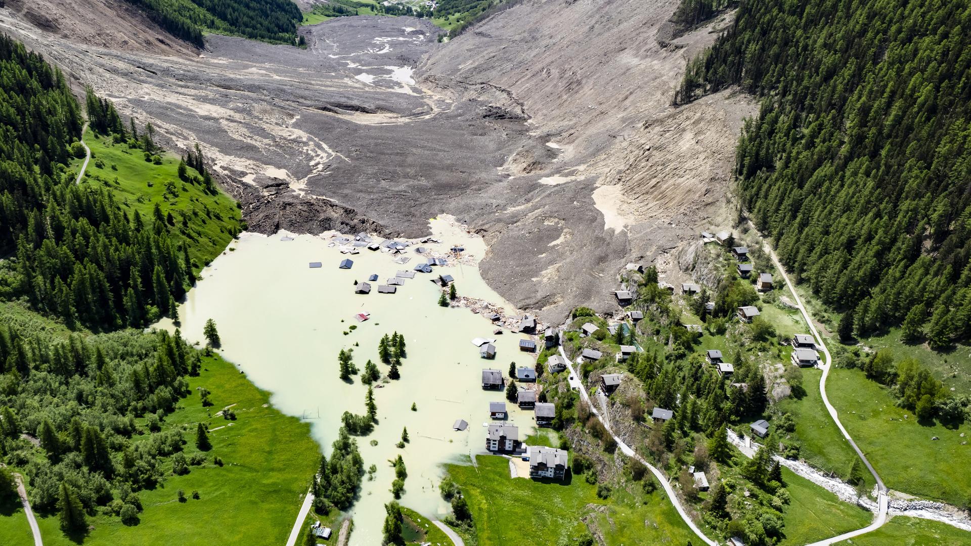 Von Berghängen ist viel Geröll heruntergekommen. Einige Häuser von einem Dorf sind verschüttet, andere Häuser stehen im Wasser. Ein See hat sich gebildet. Von Berghängen ist viel Geröll heruntergekommen. Einige Häuser von einem Dorf sind verschüttet, andere Häuser stehen im Wasser. Ein See hat sich gebildet.