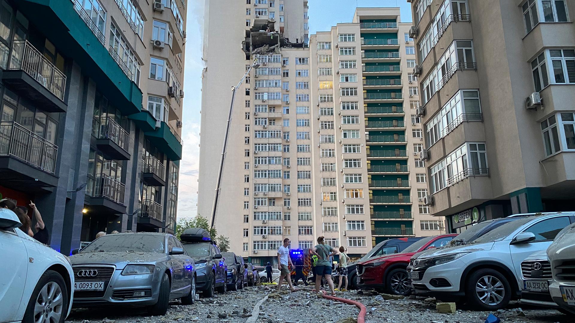 Menschen stehen in Kiew auf einem Parkplatz mit beschädigten Autos. Im Hintergrund ist ein mehrstöckiges Wohnhaus zu sehen, das bei einem Luftangriff beschädigt worden ist.