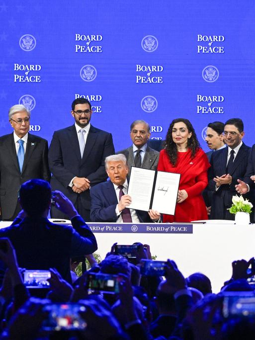 Eine Reihe an Premierministern und Prsäidenten stehen auf einer Bühne hinter Trump. Der US-Präsident hält einen unterschriebenen Vertrag in die Kameras