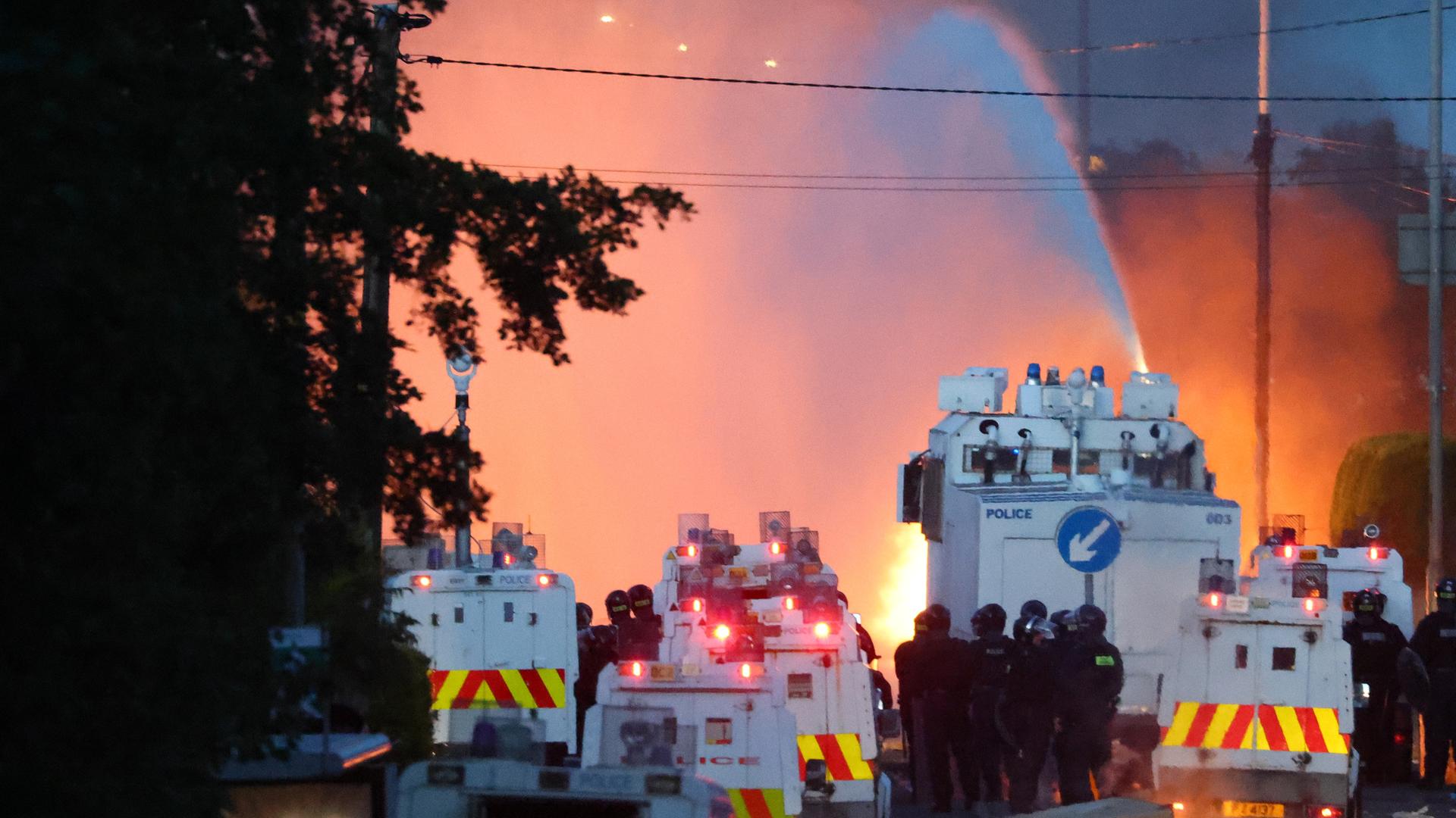 Zu sehen ist ein großes Aufgebot an Polizeikräften und Einsatzfahrzeugen. In der Nähe lodern Feuer, aus einem Wasserwerfer wird ein Wasserstrahl in eine Richtung gehalten.