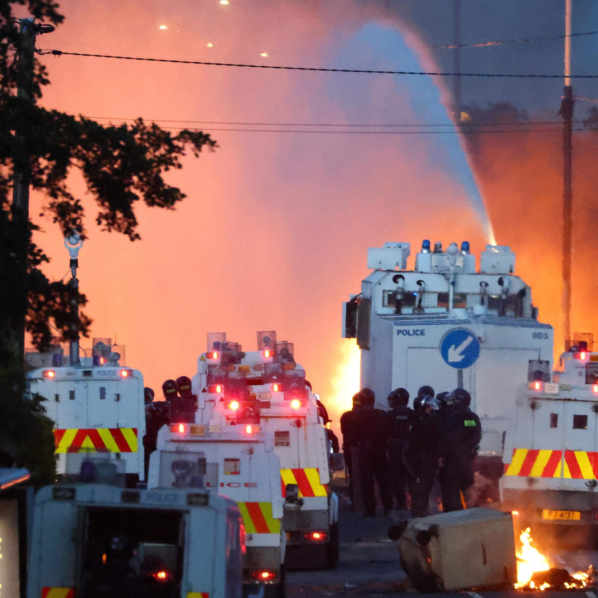 Zu sehen ist ein großes Aufgebot an Polizeikräften und Einsatzfahrzeugen. In der Nähe lodern Feuer, aus einem Wasserwerfer wird ein Wasserstrahl in eine Richtung gehalten.