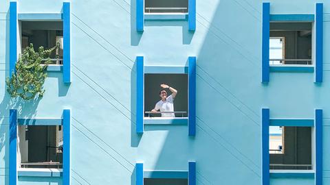 Ein Mann am Fenster eines Hauses mit blauer Fassade schützt seine Augen vor der Sonne.