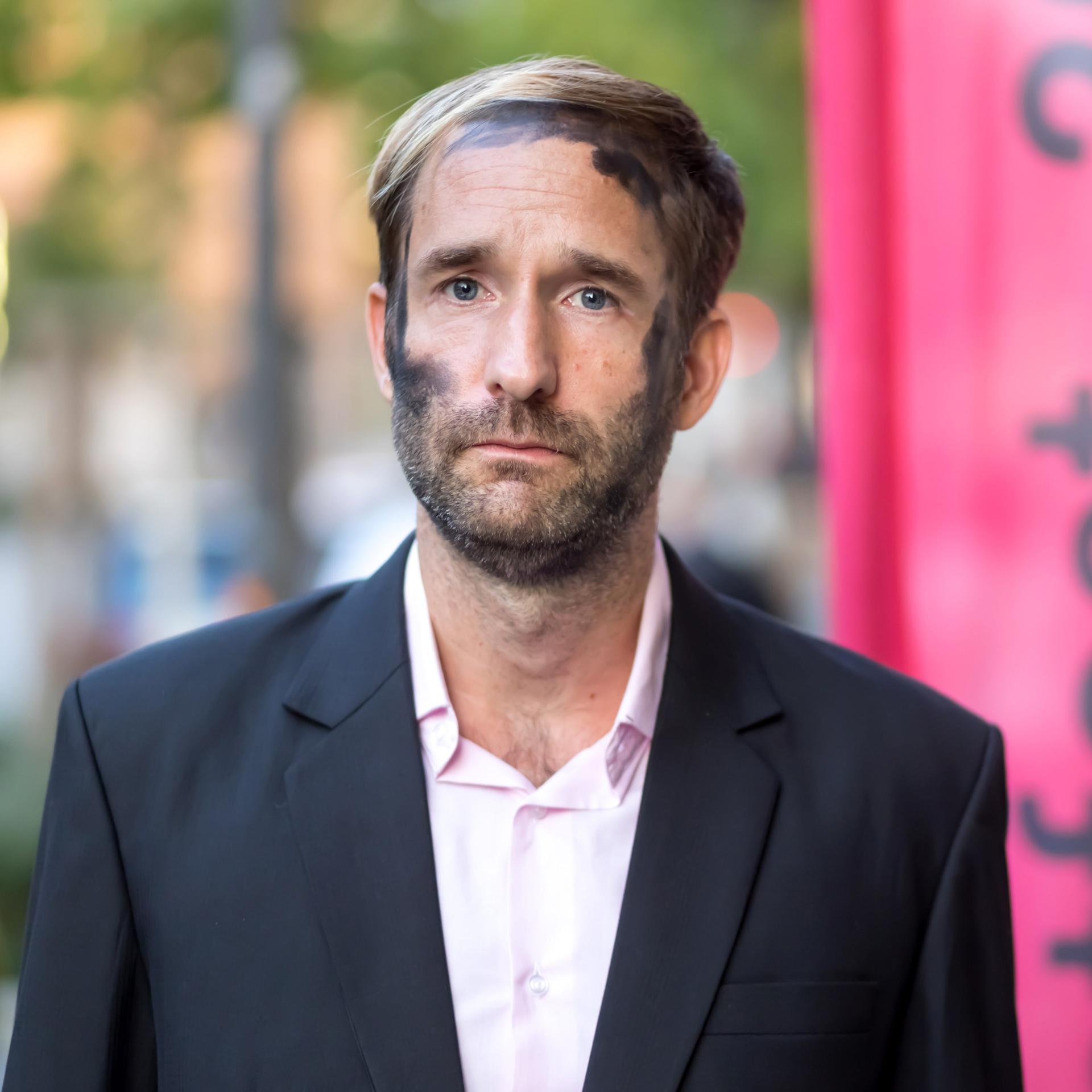 Philipp Ruch blickt ernst frontal in die Kamera. In seinem Gesicht sind die schwarzen Kohlespuren zu sehen, die er bei Aktionen des Zentrums für Politische Schönheit traditionell trägt
