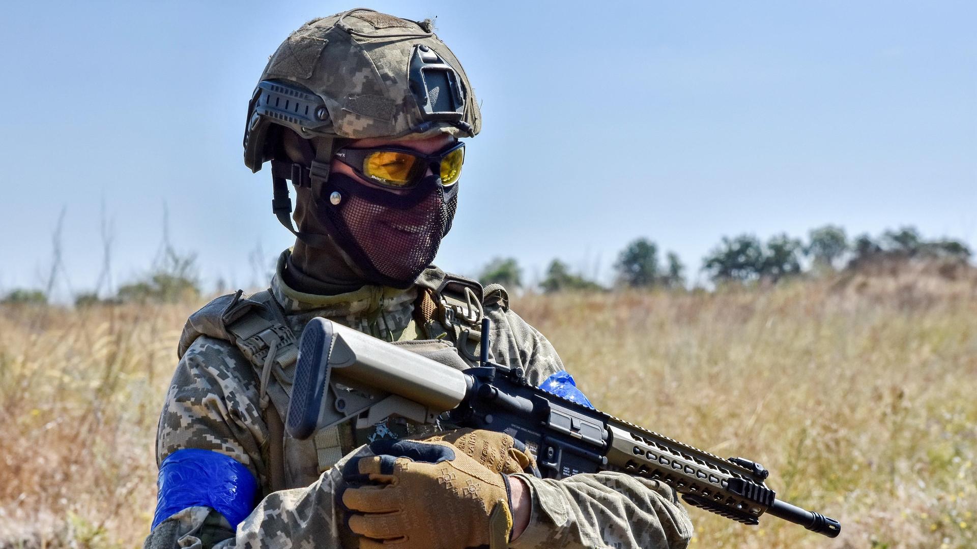 Ein ukrainischer Soldat steht in Ausrüstung mit einem Gewehr in der Hand und lächelt.