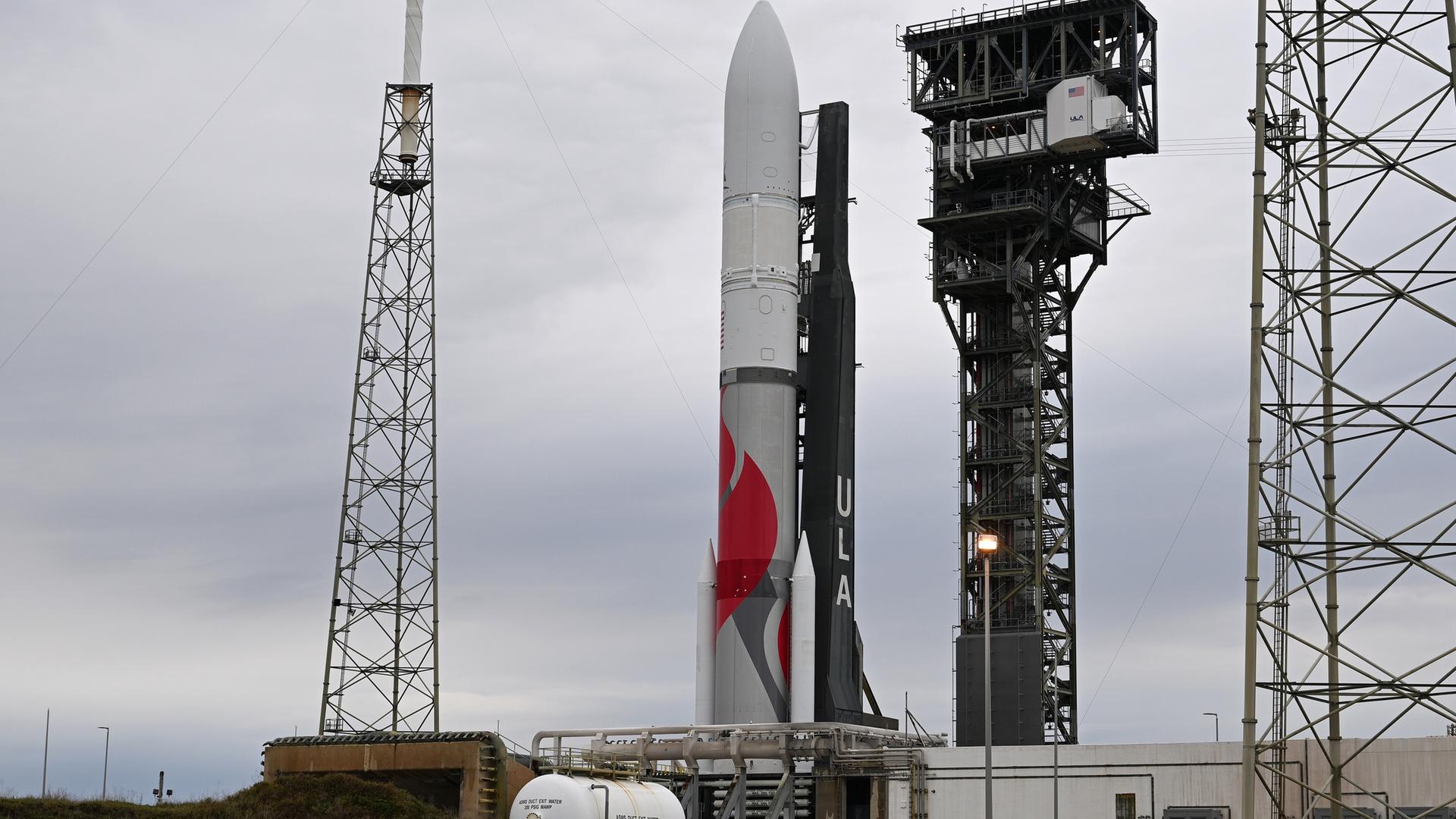 Blick auf die aufrecht stehende rot-weiße Rakete an der Startrampe. Davor zahlreiche weiße Tanks. Der Himmel ist grau. Blick auf die aufrecht stehende rot-weiße Rakete an der Startrampe. Davor zahlreiche weiße Tanks. Der Himmel ist grau.