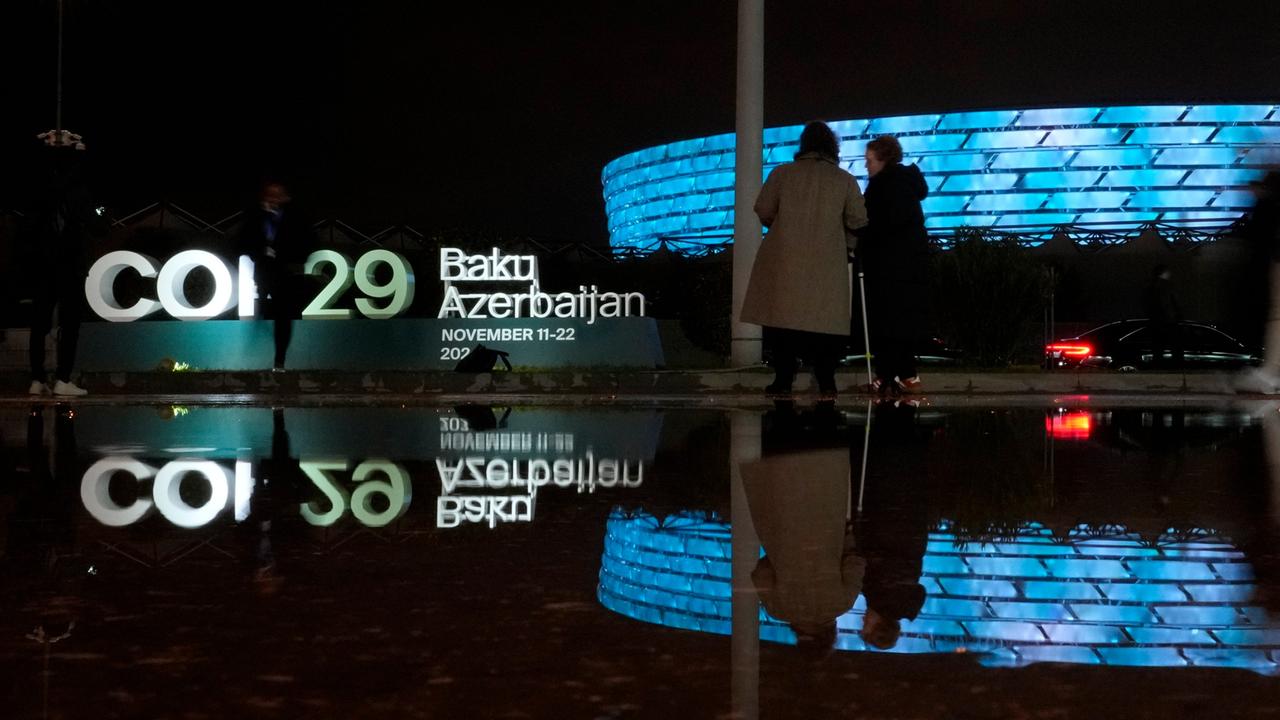 Im Vordergrund ist ein leuchtendes Logo zu sehen. Menschen stehen herum. Zwe Peronen scheinen sich zu unterhalten. Im Hintergrund ist ein großes leuchtendes Gebäude zu sehen. Die gesamte Szenerie spiegelt sich in einer dunklen Wasserfläche im Vordergrund.