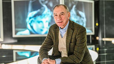 Rainer Rother sitzt im Deutsche Kinemathek Museum für Film und Fernsehen. Im Hintergrund sind Bildschirme mit Filmszenen zu sehen. Er trägt ein hellblaues Hemd, einen beigen Pullover und ein grün-kariertes Sakko.