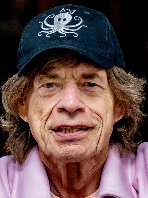 Porträtaufnahme von Mick Jagger, der freundlich grinst. Er trägt Hut und einen rosa Pulli.