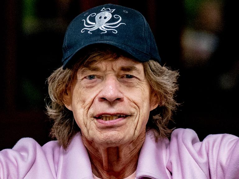 Porträtaufnahme von Mick Jagger, der freundlich grinst. Er trägt Hut und einen rosa Pulli.