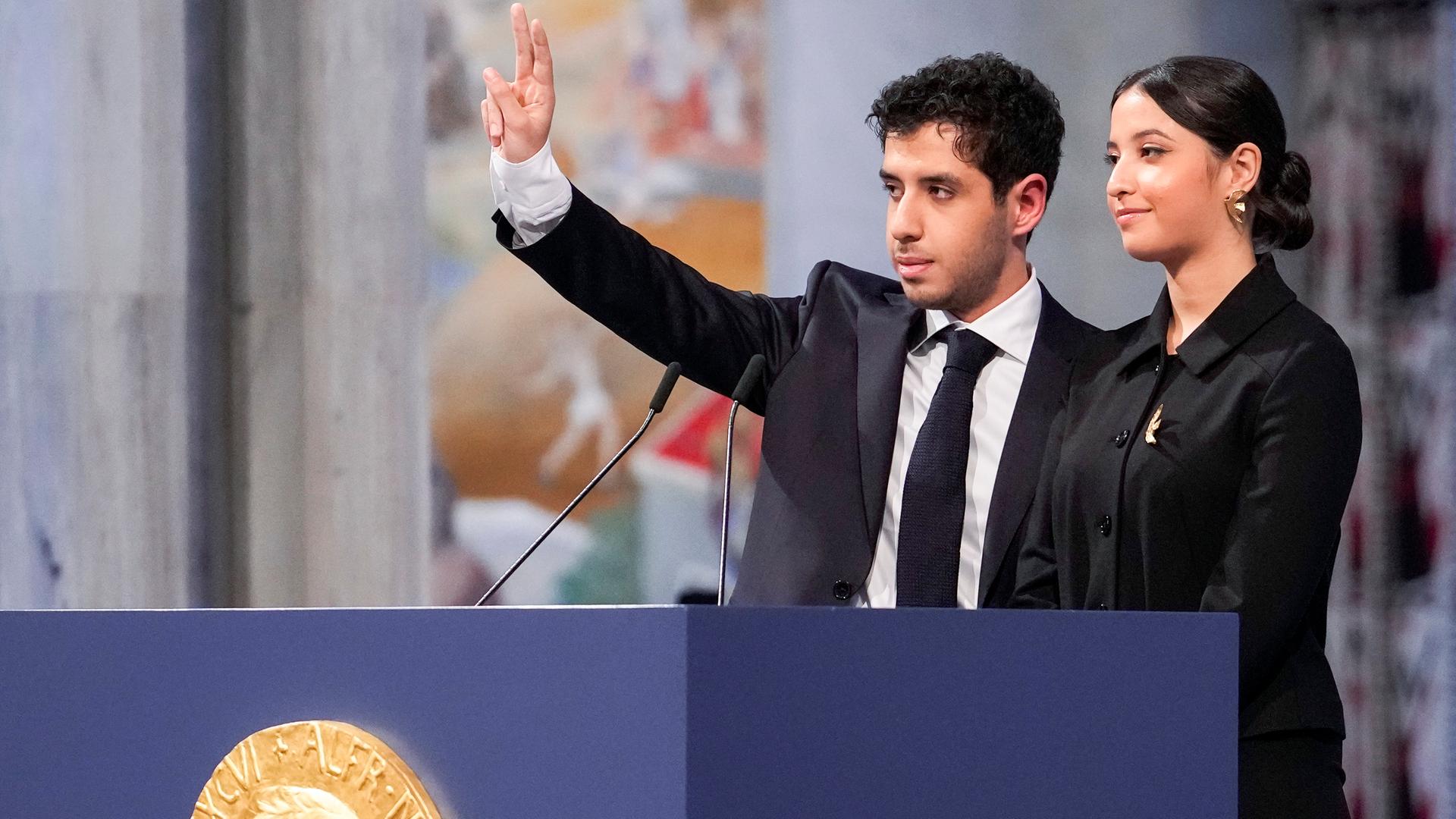 Der junge Mann steht neben seiner jungen Schwester, beide sind festlich gekleidet. Er grüßt mit der rechten Hand. Vor ihnen das Rednerpult mit einer großen Nachbildung der Medaille und dem Konterfei von Alfred Nobel Der junge Mann steht neben seiner jungen Schwester, beide sind festlich gekleidet. Er grüßt mit der rechten Hand. Vor ihnen das Rednerpult mit einer großen Nachbildung der Medaille und dem Konterfei von Alfred Nobel
