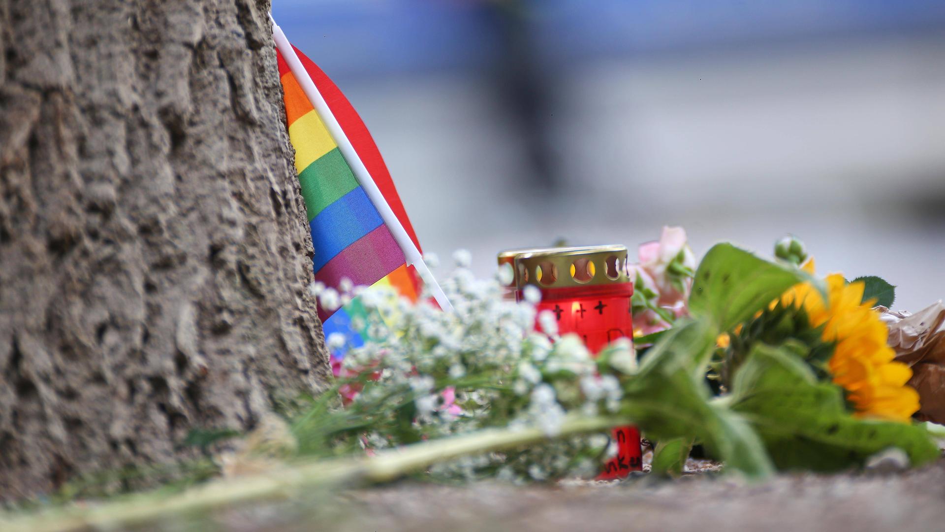 An einem Baumstamm lehnt eine kleine Regenbogenflagge. Davor liegen Blumen und stehen Kerzen. An einem Baumstamm lehnt eine kleine Regenbogenflagge. Davor liegen Blumen und stehen Kerzen.