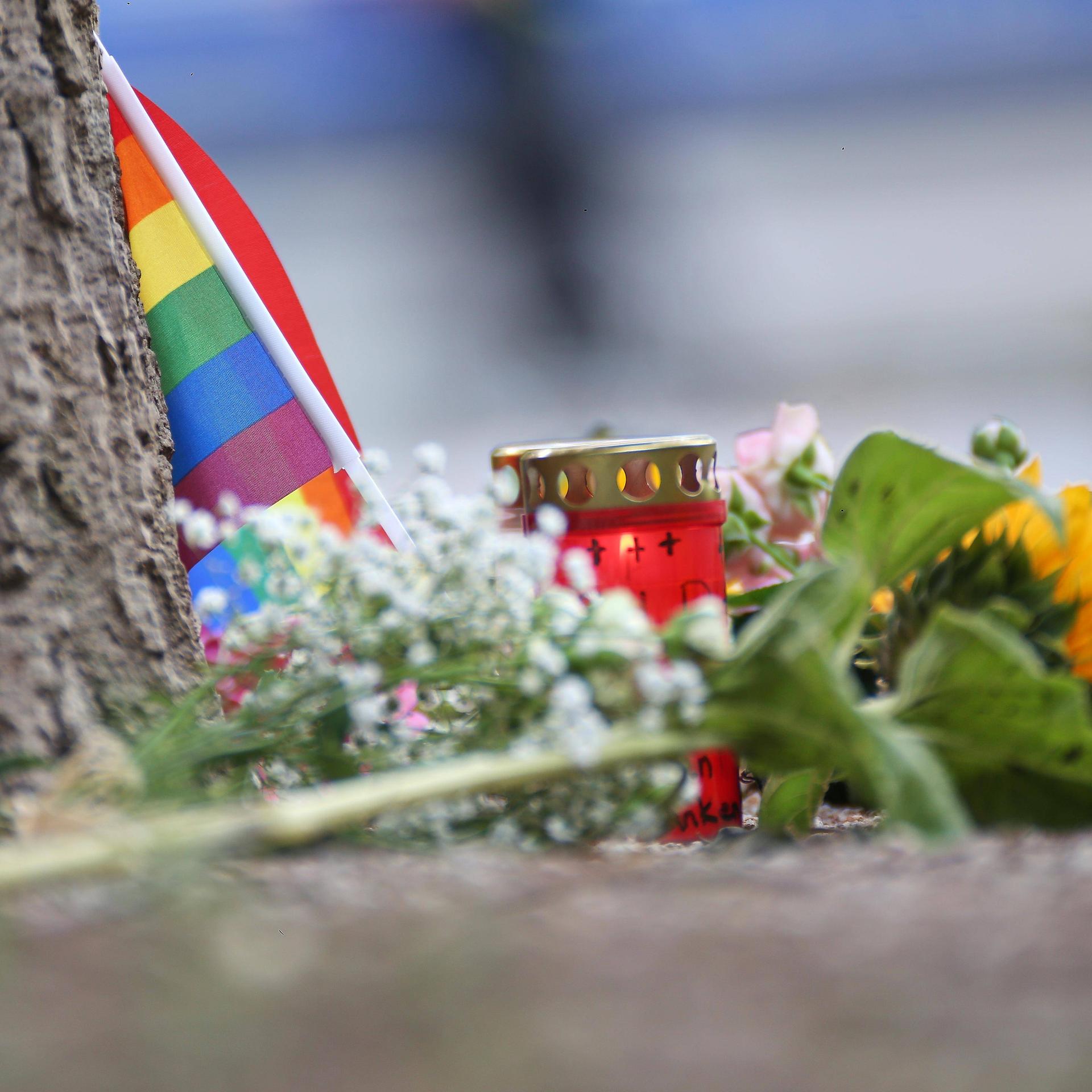 An einem Baumstamm lehnt eine kleine Regenbogenflagge. Davor liegen Blumen und stehen Kerzen.