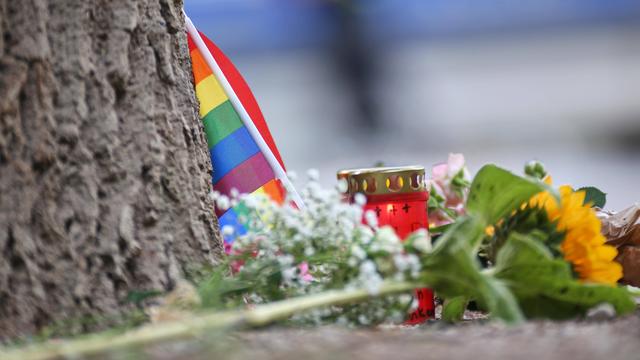 An einem Baumstamm lehnt eine kleine Regenbogenflagge. Davor liegen Blumen und stehen Kerzen.