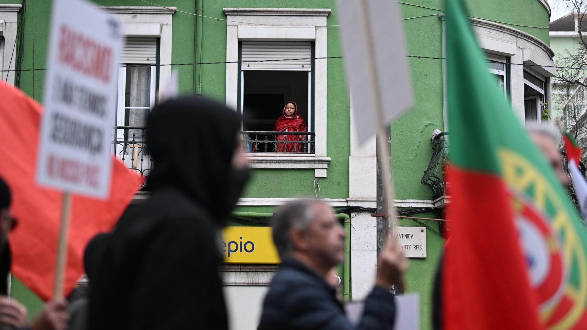 Demonstranten ziehen mit Plakaten und portugiesischen Flaggen eine Straße entlang. Im Hintergrund schaut eine Frau aus einem Fenster im ersten Stock hinab auf den Protest.