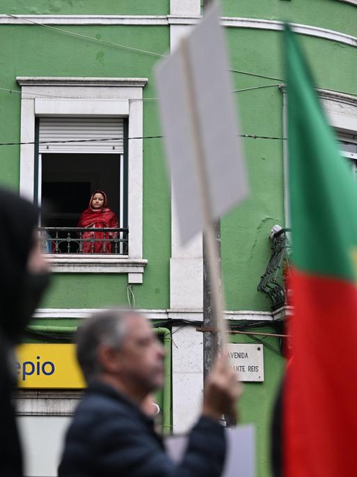 Demonstranten ziehen mit Plakaten und portugiesischen Flaggen eine Straße entlang. Im Hintergrund schaut eine Frau aus einem Fenster im ersten Stock hinab auf den Protest.