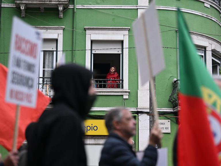 Demonstranten ziehen mit Plakaten und portugiesischen Flaggen eine Straße entlang. Im Hintergrund schaut eine Frau aus einem Fenster im ersten Stock hinab auf den Protest.