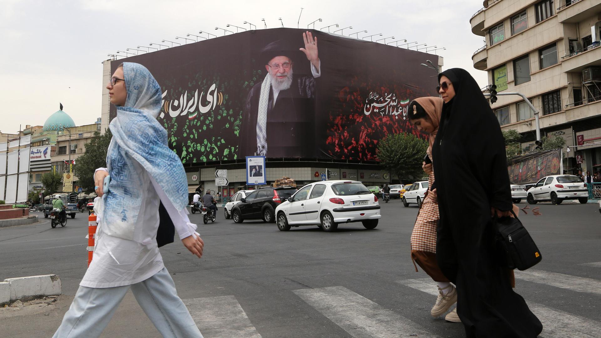 Kopftuch tragende Frauen überqueren eine Straße, im Hintergrund ein großes Bild von Ayatollah Ali Khamenei an einer Häuserwand