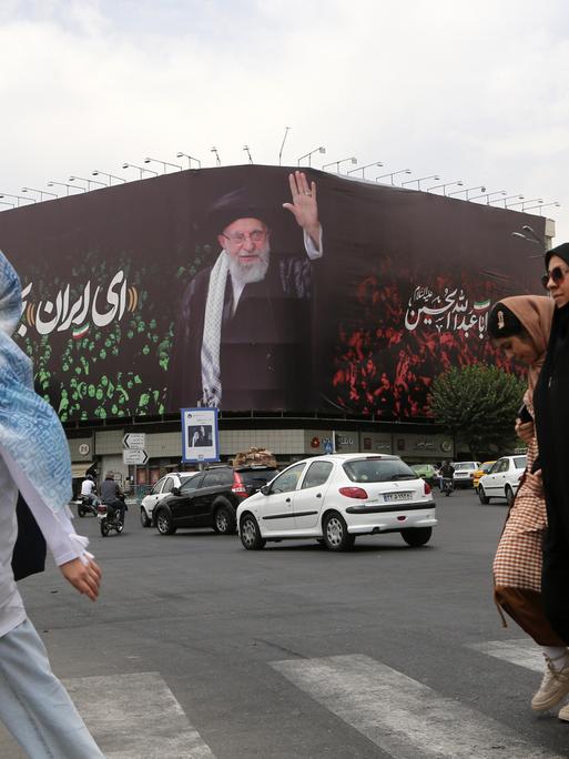 Kopftuch tragende Frauen überqueren eine Straße, im Hintergrund ein großes Bild von Ayatollah Ali Khamenei an einer Häuserwand