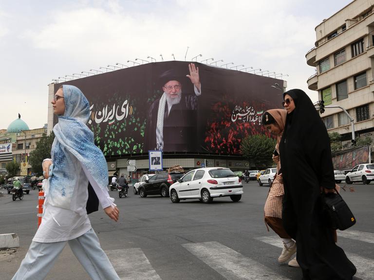 Kopftuch tragende Frauen überqueren eine Straße, im Hintergrund ein großes Bild von Ayatollah Ali Khamenei an einer Häuserwand