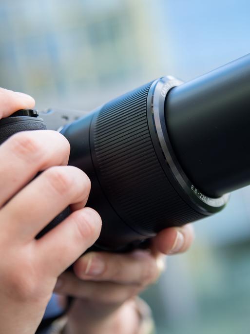 Seitliche Großaufnahme einer Fotokamera, die von zwei Händen gehalten wird. Der rechte Zeigefinger liegt auf dem Auslöser. 
