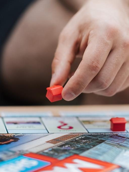 Eine Hand versetzt ein Spielhaus auf einem Monopoly-Spielbrett. Eine Hand versetzt ein Spielhaus auf einem Monopoly-Spielbrett.