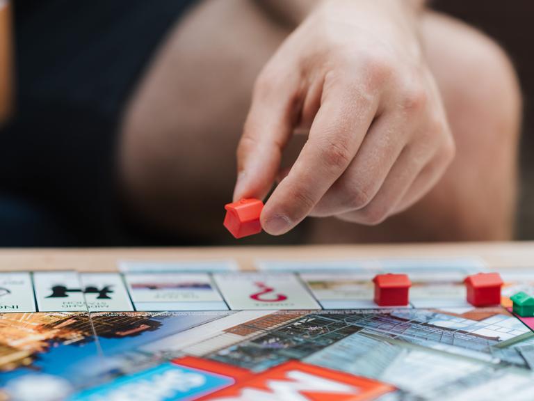 Eine Hand versetzt ein Spielhaus auf einem Monopoly-Spielbrett. Eine Hand versetzt ein Spielhaus auf einem Monopoly-Spielbrett.