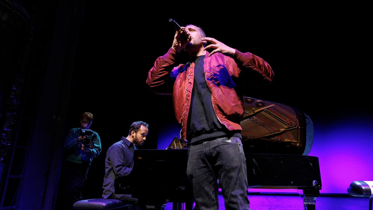 Danger Dan singt stehend in ein Mikrofon. Im Hintergrund sitzt Igor Levit am Flügel und begleitet ihn. 