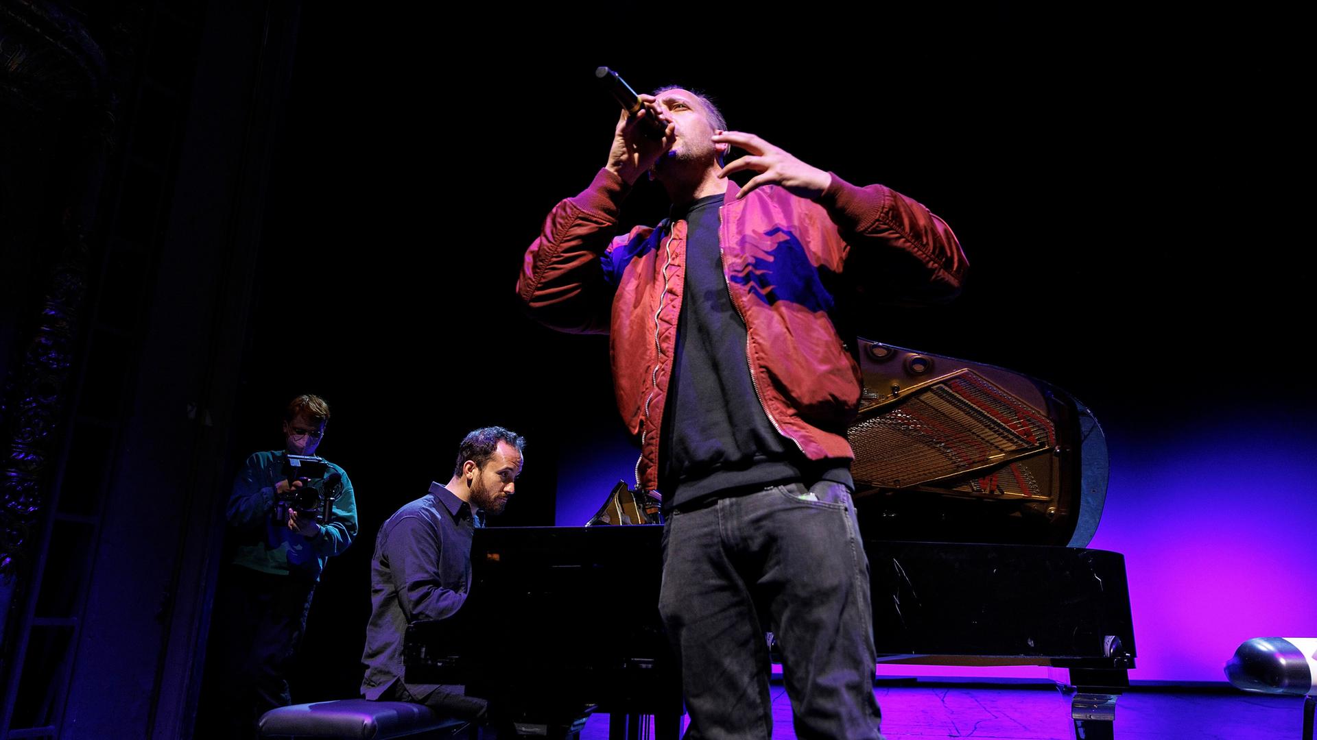Danger Dan singt stehend in ein Mikrofon. Im Hintergrund sitzt Igor Levit am Flügel und begleitet ihn. Danger Dan singt stehend in ein Mikrofon. Im Hintergrund sitzt Igor Levit am Flügel und begleitet ihn.
