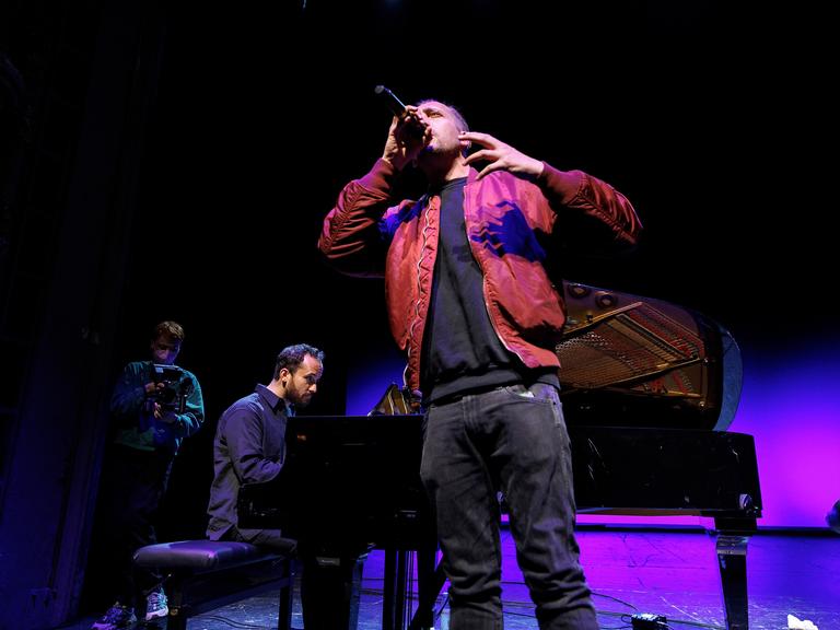 Danger Dan singt stehend in ein Mikrofon. Im Hintergrund sitzt Igor Levit am Flügel und begleitet ihn. 