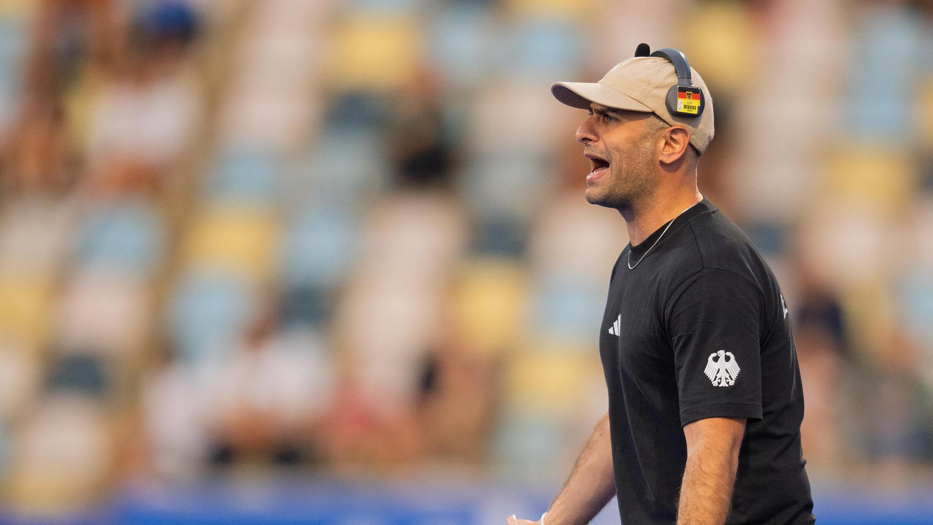 Hockey-Bundestrainer Henning ruft etwas vom Spielfeld-Rand. Er hat ein schwarzes Trikot an und eine beige Kappe auf und trägt darüber einen Kopfhörer mit Deutschland-Farben. 