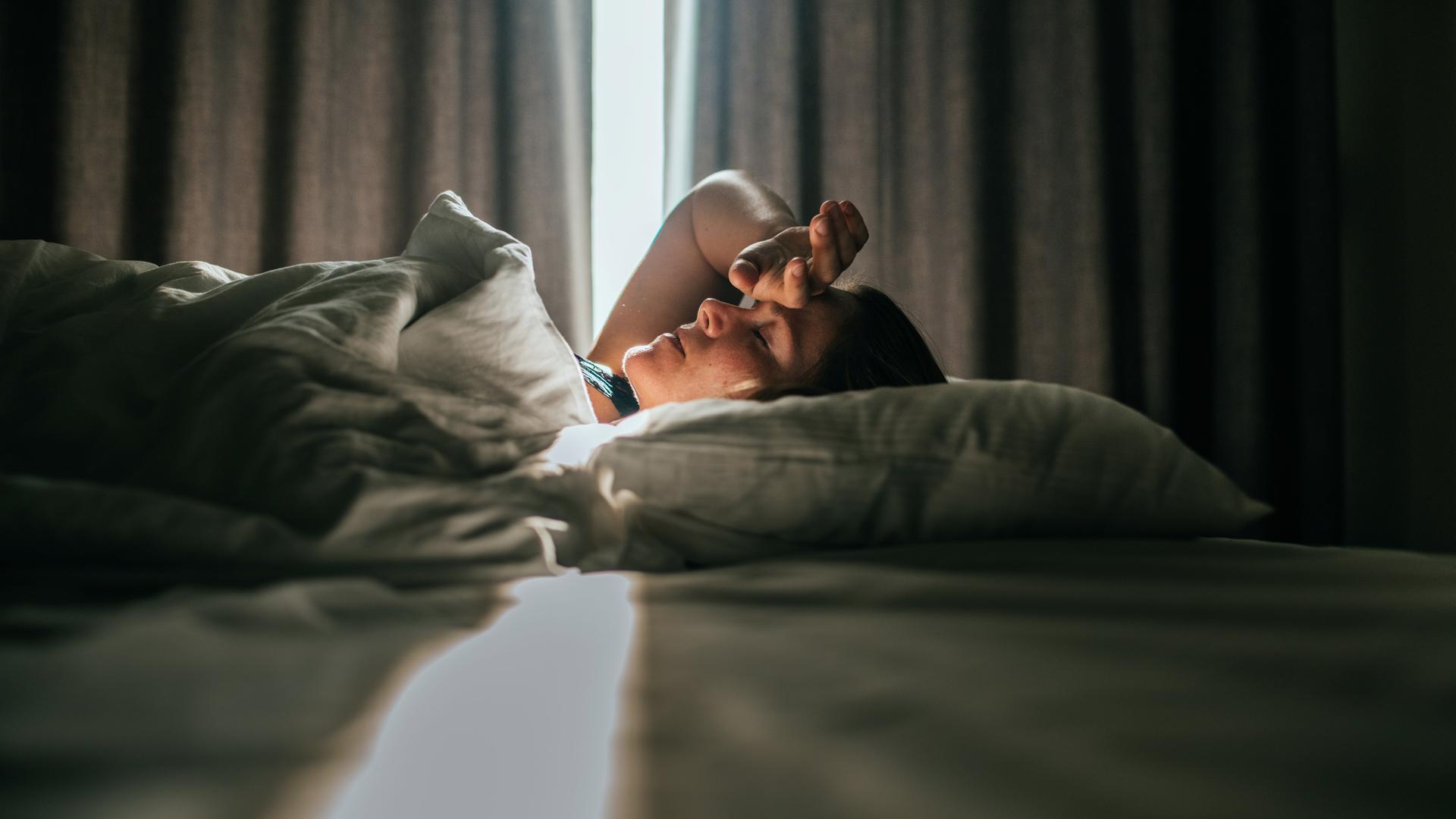 Eine Frau liegt im Bett. Sonniges Tageslicht fällt durch die Vorhänge im Hintergrund. Sie hatte eine Hand an die Stirn gelegt.