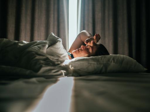 Eine Frau liegt im Bett. Sonniges Tageslicht fällt durch die Vorhänge im Hintergrund. Sie hatte eine Hand an die Stirn gelegt. Eine Frau liegt im Bett. Sonniges Tageslicht fällt durch die Vorhänge im Hintergrund. Sie hatte eine Hand an die Stirn gelegt.